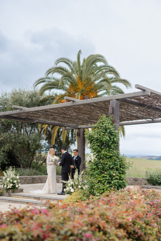 Wedding ceremony at Carneros Resort and Spa with the couple exchanging vows beneath the arbor, framed by palm trees and vineyard views in Napa Valley