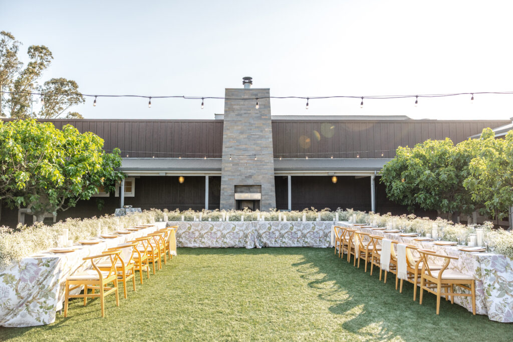 Outdoor wedding reception at Carneros Resort and Spa featuring long banquet tables on the lawn, wooden chairs, lush greenery, and the signature stone fireplace in Napa Valley