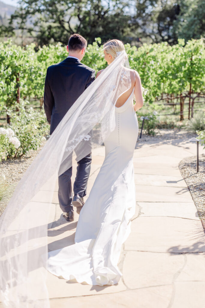 Newly married couple walking hand in hand through the vineyard paths at Estate Yountville, with the bride’s veil flowing behind her in Napa Valley.