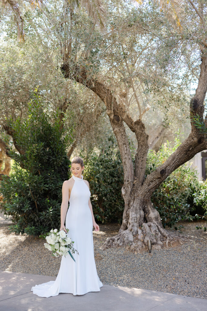 Bride holding a white bouquet while standing beneath olive trees at Estate Yountville, photographed in natural Napa Valley light.