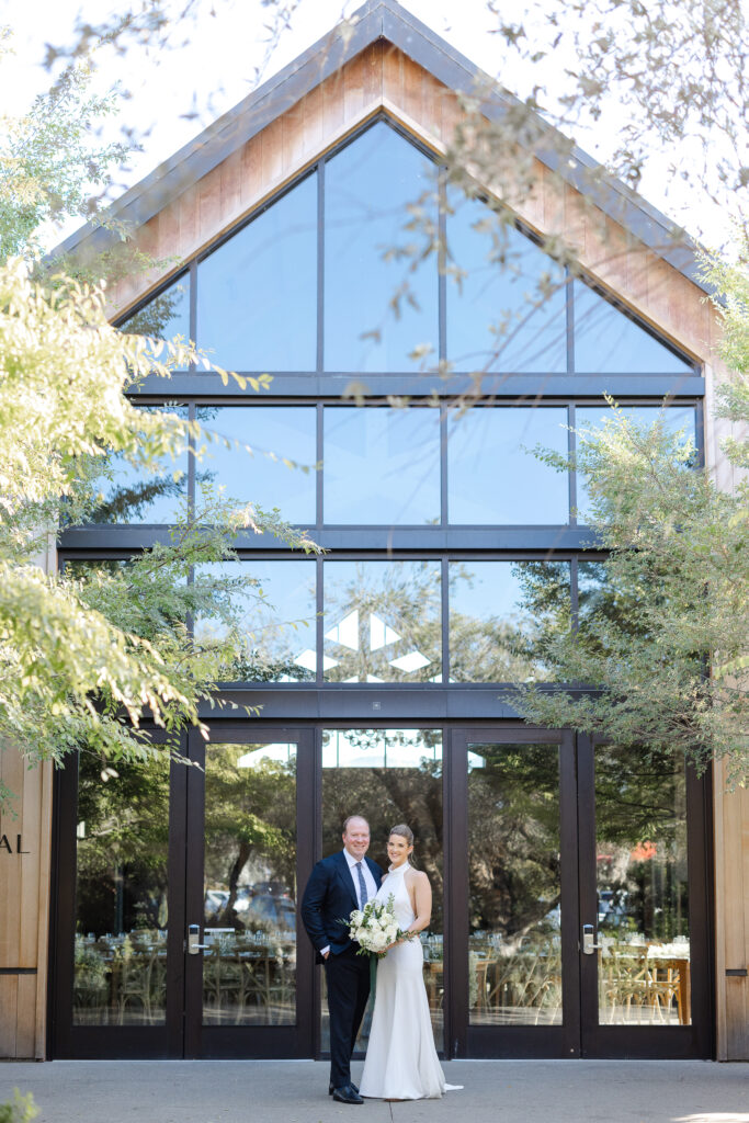 Bride and groom standing together outside the Estate Yountville reception building with modern wood architecture and glass doors.