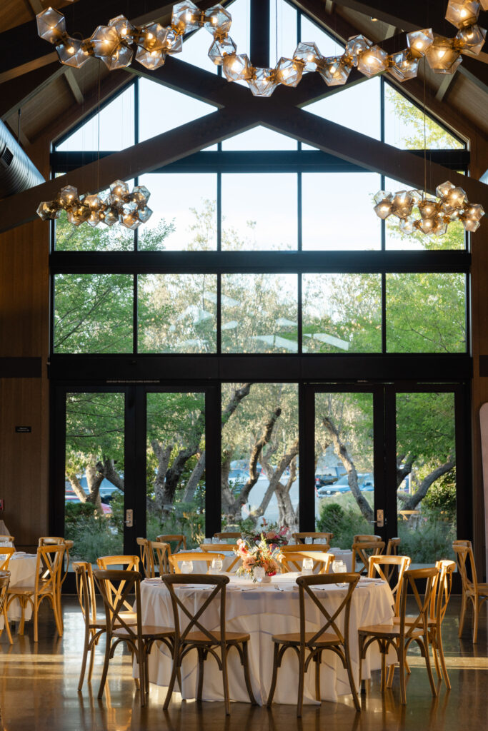 Elegant wedding reception inside the Estate Yountville pavilion featuring long wooden tables, white linens, modern chandeliers, and floor-to-ceiling windows overlooking Napa Valley greenery.