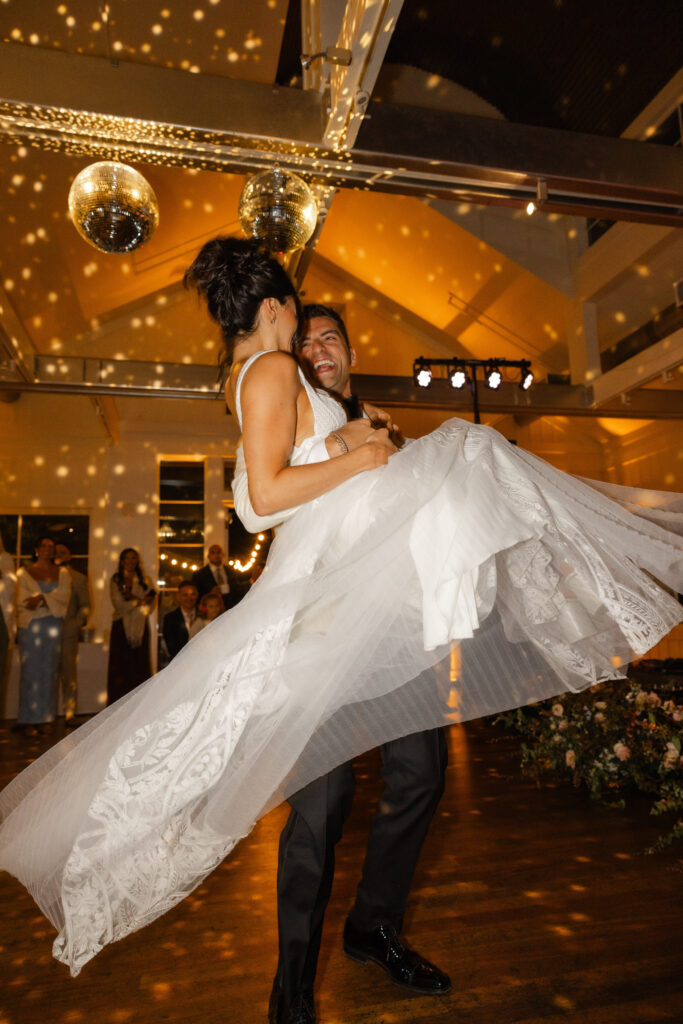 Groom lifting the bride mid dance during their wedding reception, surrounded by warm disco ball lighting and cheering guests at Carneros Resort and Spa in Napa Valley.