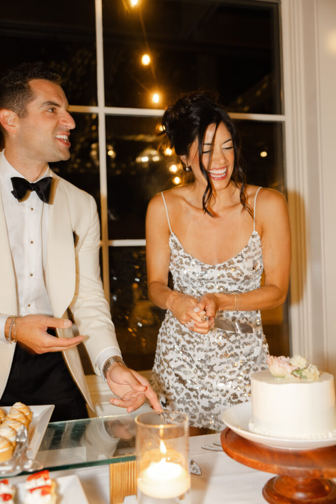 Bride and groom laughing together while cutting their wedding cake during the reception, candlelight glowing around the dessert table at Carneros Resort and Spa.