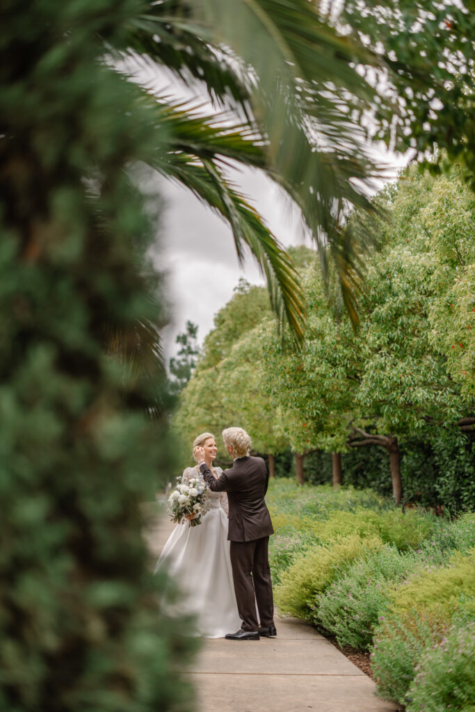 Bride and groom sharing an intimate moment along a garden path at Carneros Resort and Spa, framed by lush greenery as the groom gently adjusts the bride’s hair while she holds a white floral bouquet.