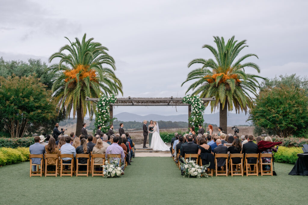 Wedding ceremony at the arbor at Carneros Resort and Spa with the couple exchanging vows beneath a floral arch framed by palm trees and vineyard views