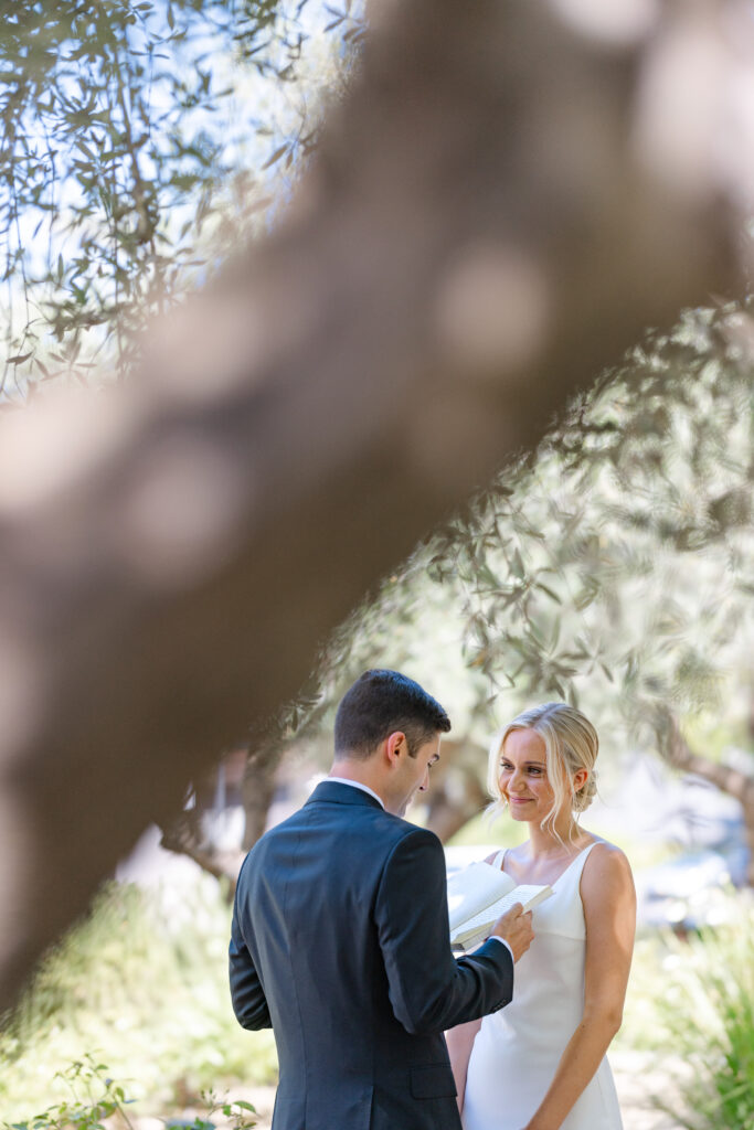 Bride and groom sharing a quiet vow reading beneath olive trees at Estate Yountville, framed by soft greenery and filtered Napa Valley light.
