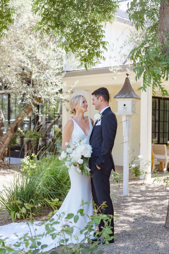 Bride and groom standing together in the garden courtyard at Estate Yountville, framed by olive trees, soft greenery, and the property’s classic white architecture.