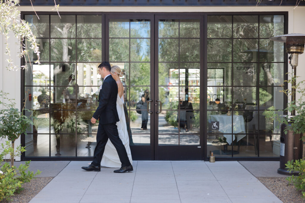 Bride and groom strolling through the garden walkway at Estate Yountville, with reflections of greenery in the glass doors behind them.