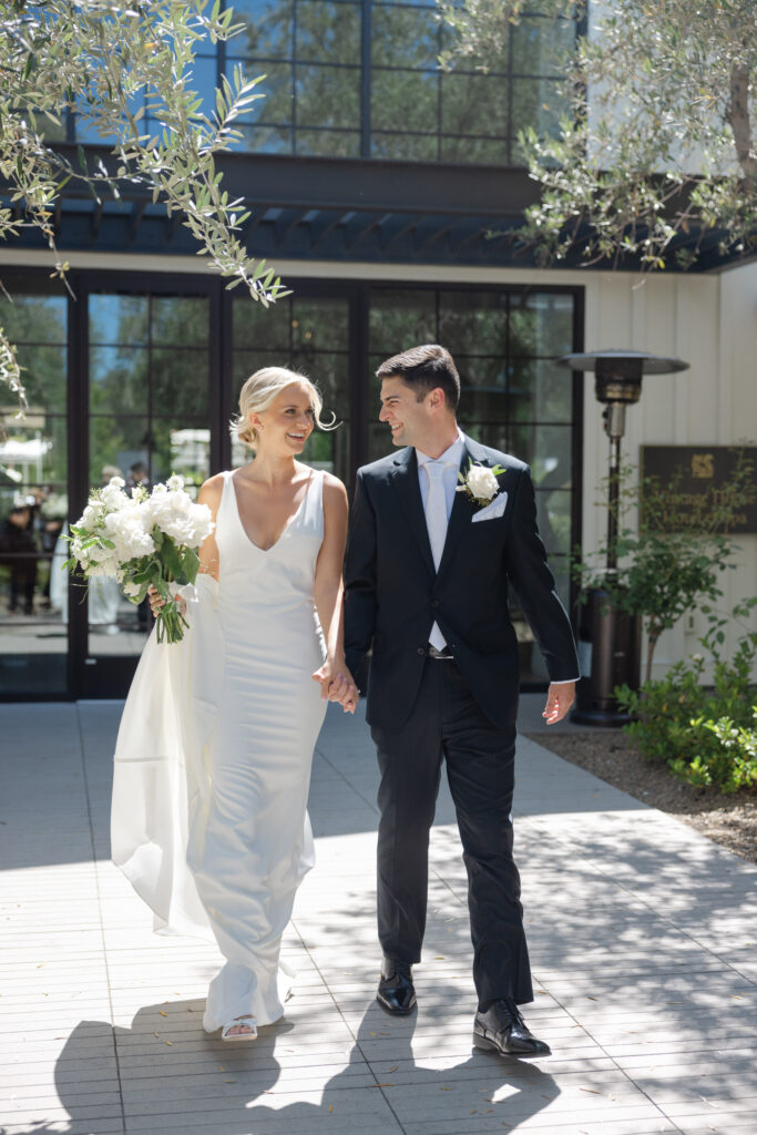 Bride and groom walking hand in hand outside The Social at Estate Yountville, captured in bright Napa Valley light with modern black-framed doors behind them.