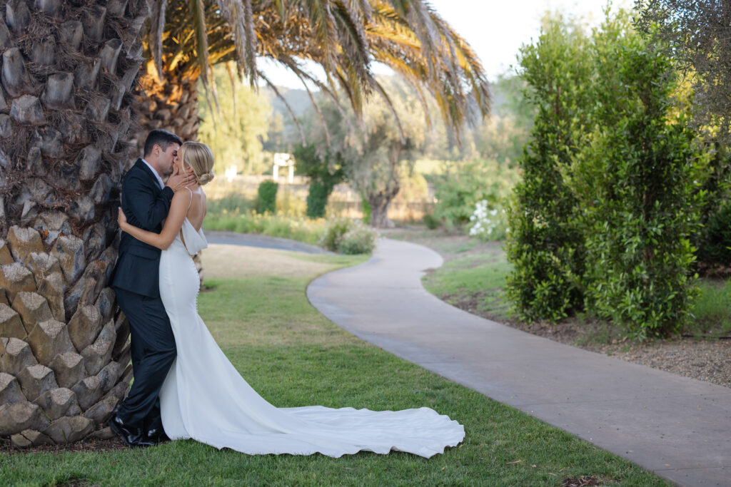 Bride and groom sharing a quiet moment beneath a palm tree at The Social at Estate Yountville, surrounded by gardens and winding pathways