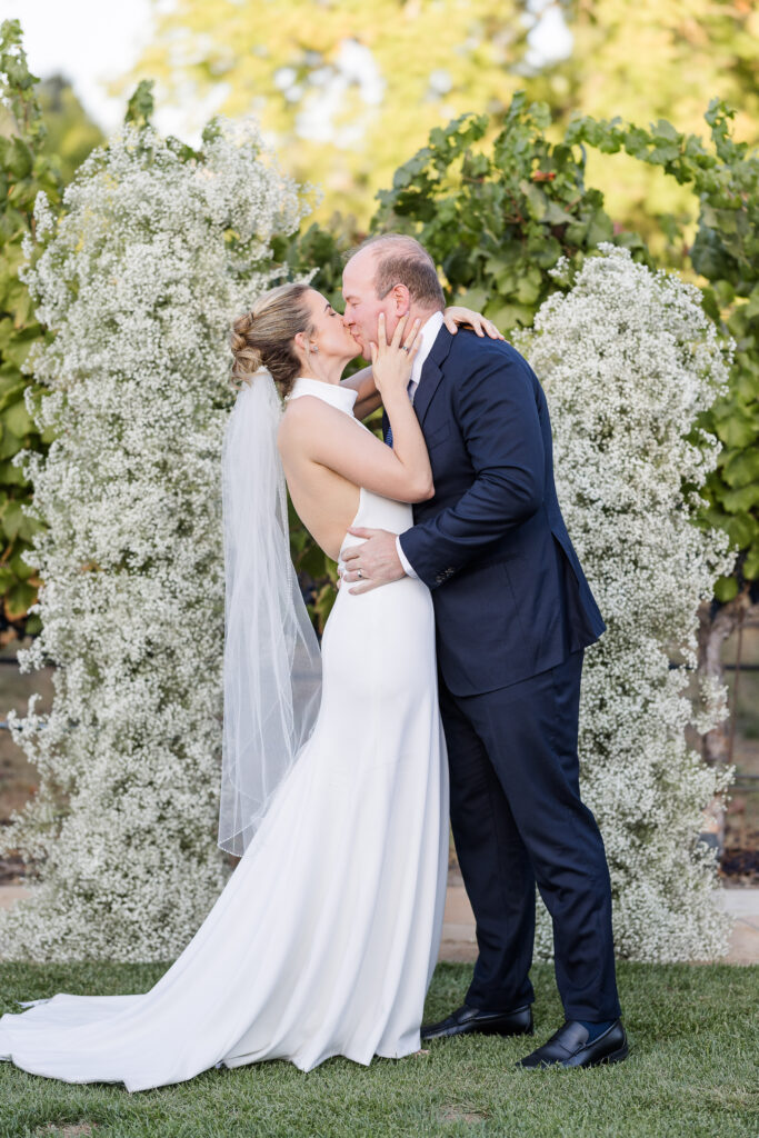 Bride and groom sharing first kiss moment during their Estate Yountville vineyard wedding, surrounded by lush vines and white florals