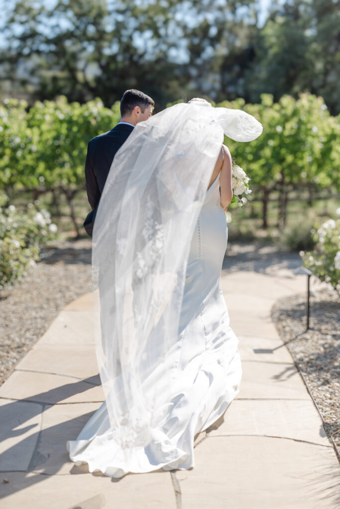 Bride and groom walking through the vineyard at Estate Yountville, bride’s veil flowing in the sunlight during a Napa Valley wedding