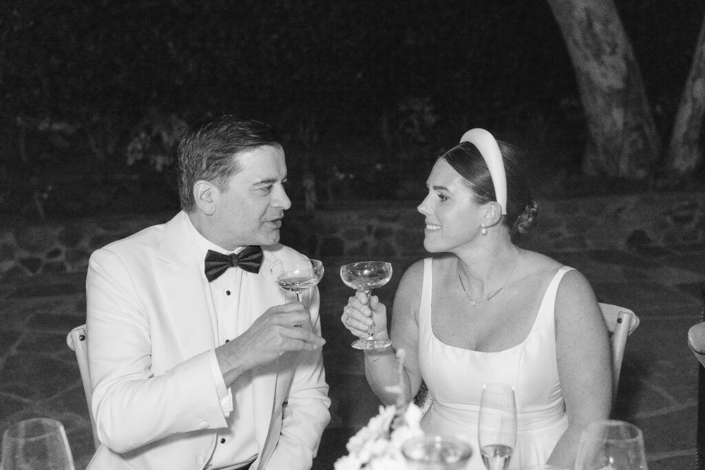 A bride and groom share a champagne toast at their Napa Valley wedding reception, the bride wearing a sleek white gown and headband while the groom looks on in a white tuxedo jacket, photographed by Napa wedding photographer Suzanne Karp