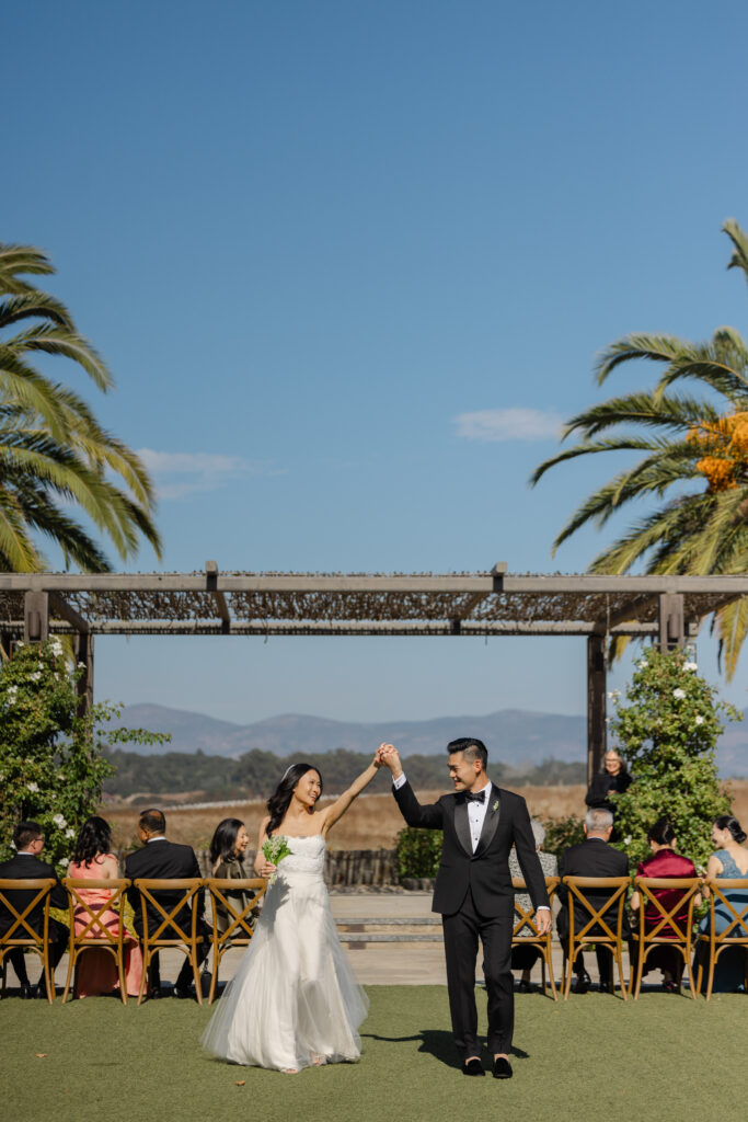 A bride and groom celebrate their recessional beneath the pergola at Carneros Resort and Spa with the Napa Valley mountains and palm trees behind them and their guests cheering, photographed by Napa wedding photographer Suzanne Karp