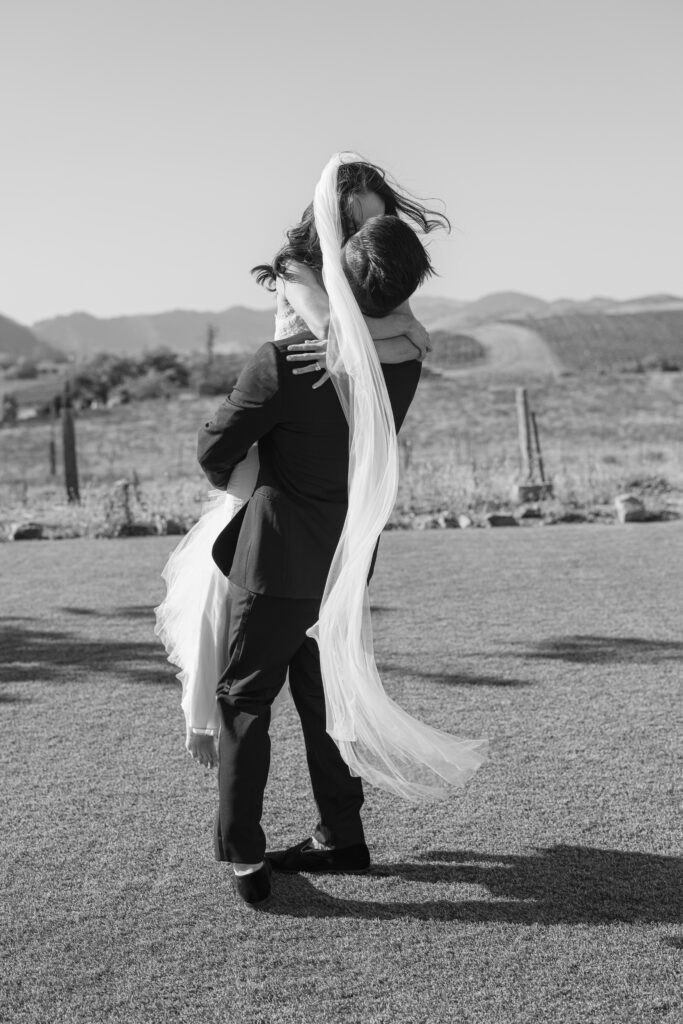 A groom lifts his bride off the ground for a kiss during their Carneros Resort elopement portraits, her veil and gown flowing in the breeze with the Napa Valley hills stretching out behind them, photographed by Napa wedding photographer Suzanne Karp