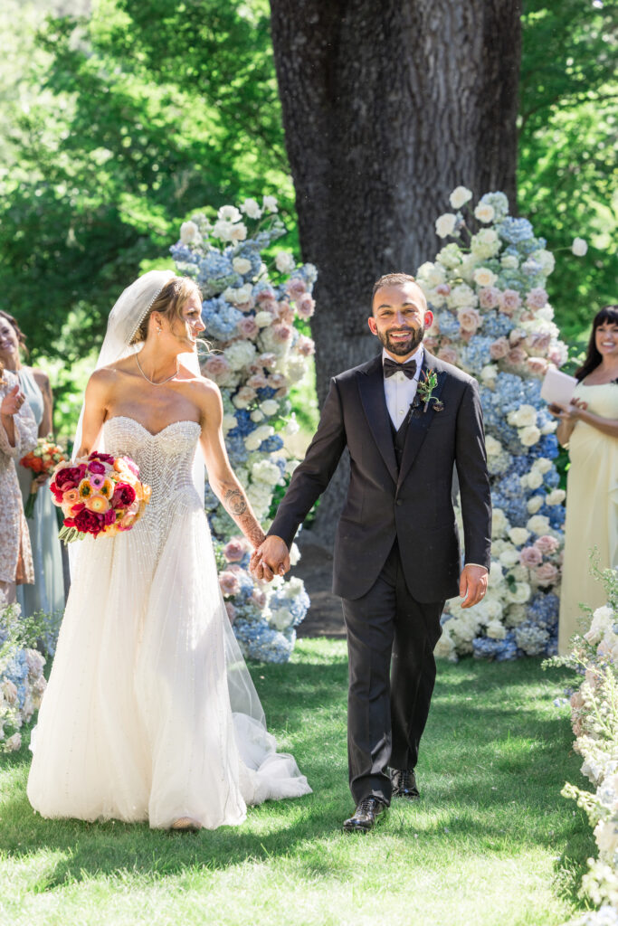A beaming bride and groom walk hand in hand down the floral lined aisle during their wedding recessional at Beaulieu Gardens in Rutherford, Napa Valley, photographed by Napa Valley wedding photographer Suzanne Karp