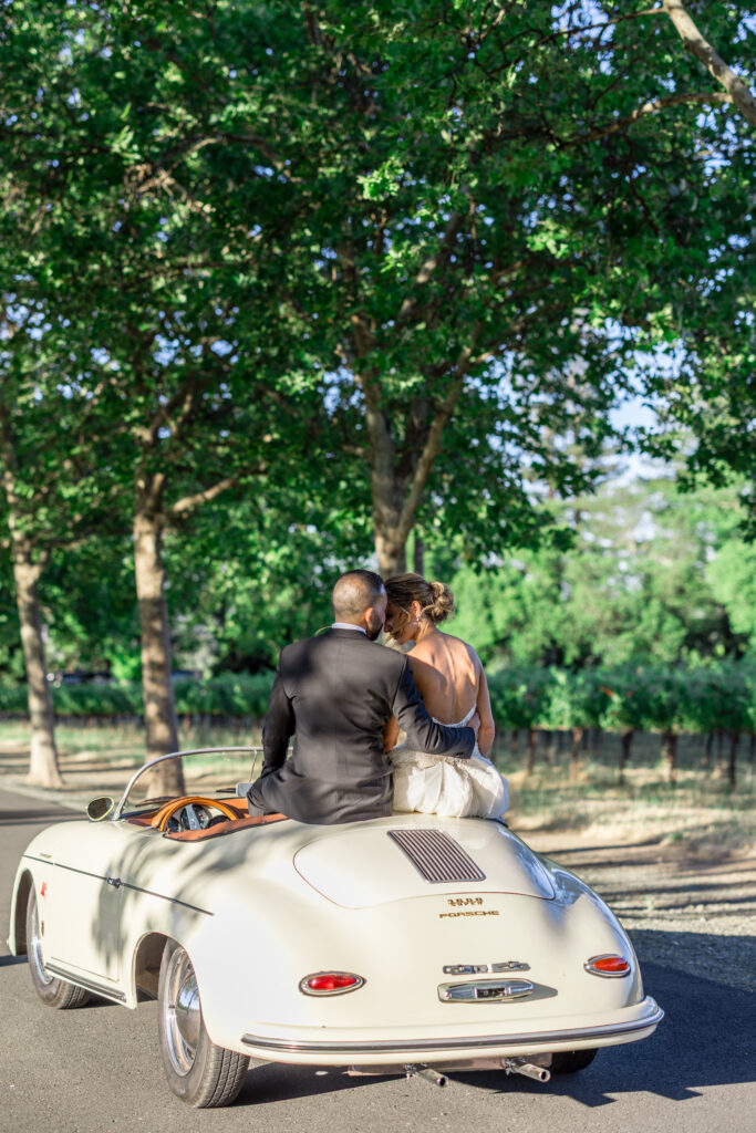 A bride and groom share a kiss in a vintage white Porsche convertible surrounded by vineyard trees during their Napa Valley wedding, photographed by Suzanne Karp Photography