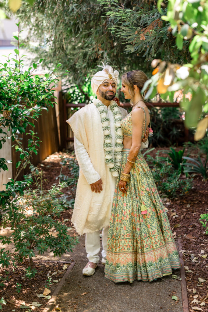 A bride and groom share a quiet moment together in a lush garden after their Hindu wedding ceremony, the bride in a sage floral printed lehenga and the groom in a cream sherwani with a varmala garland, photographed by Napa Valley wedding photographer Suzanne Karp
