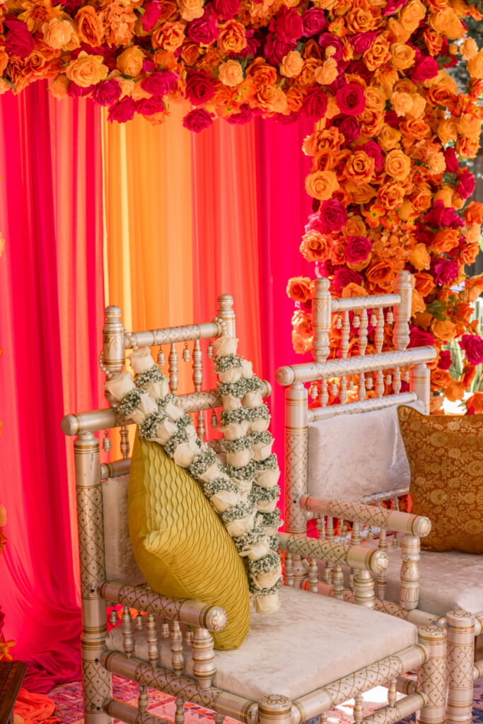 Gilded ceremonial chairs draped with white rose and baby's breath garlands sit before a wall of orange and fuchsia blooms at a Hindu wedding ceremony, photographed by Napa Valley wedding photographer Suzanne Karp