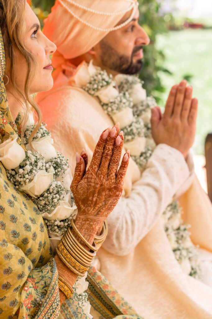 A bride and groom sit side by side in prayer during their Hindu wedding ceremony, the bride's henna covered hands and gold bangles pressed together in namaste alongside the groom's hands, photographed by Suzanne Karp Photography