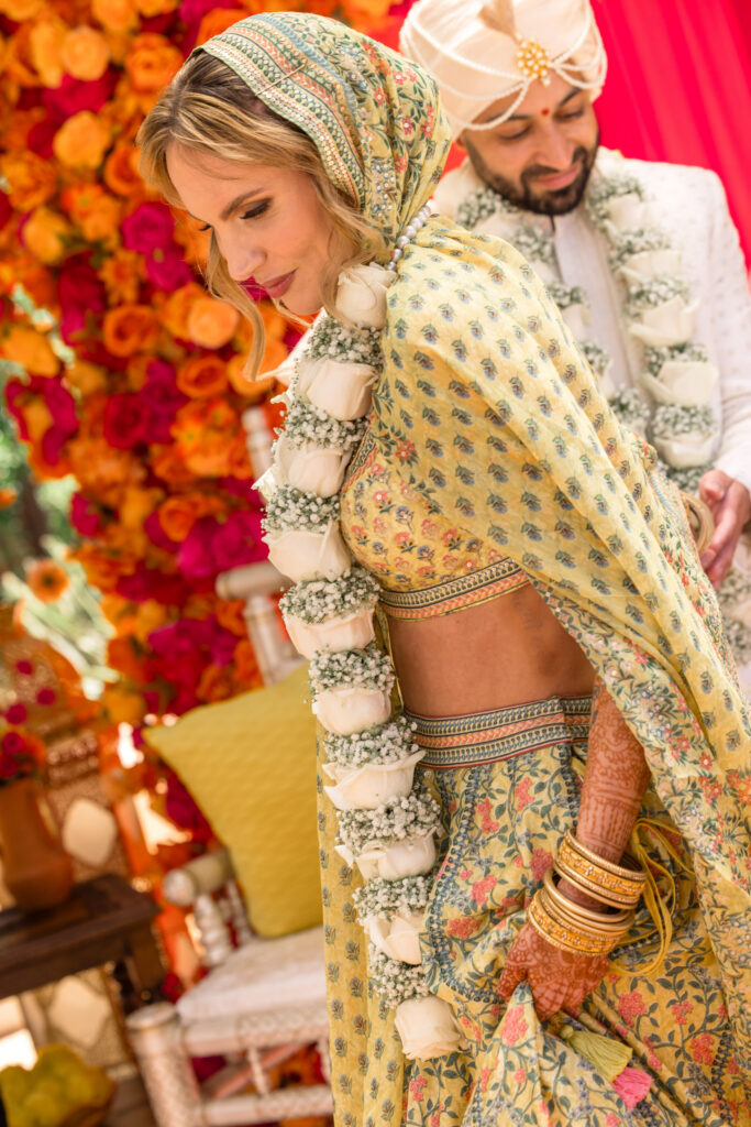 A bride in a sage printed lehenga bows her head during the varmala exchange at her Hindu wedding ceremony, her henna covered hands and gold bangles visible against a backdrop of orange and fuchsia florals, photographed by Napa Valley wedding photographer Suzanne Karp