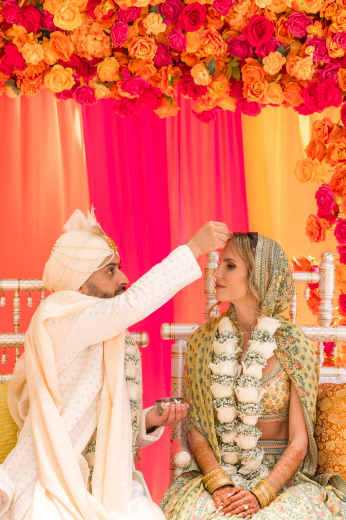 A groom in a cream sherwani and white turban applies sindoor to his bride during their Hindu wedding ceremony, the bride seated in a sage printed lehenga before a wall of orange and fuchsia roses, photographed by Suzanne Karp Photography