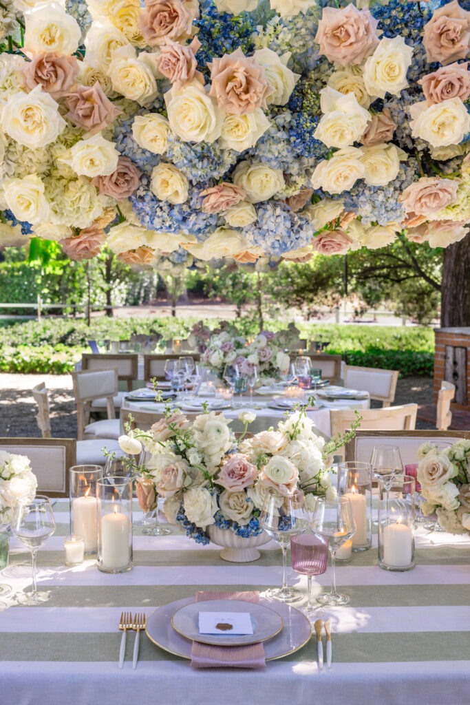 A wide shot of the reception dinner tables beneath the fig tree trellis at Beaulieu Gardens in Rutherford, set with sage stripe linens, blush and cream floral centerpieces, mixed china, and colored glassware during a Napa Valley wedding, photographed by Suzanne Karp Photography