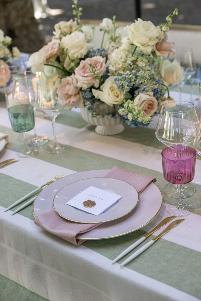 An elegant wedding place setting detail at Beaulieu Gardens featuring a blush charger plate, pink linen napkin with a gold wax seal place card, blush rose and cream centerpiece, and colored glassware on a sage stripe linen table, photographed by Suzanne Karp Photography