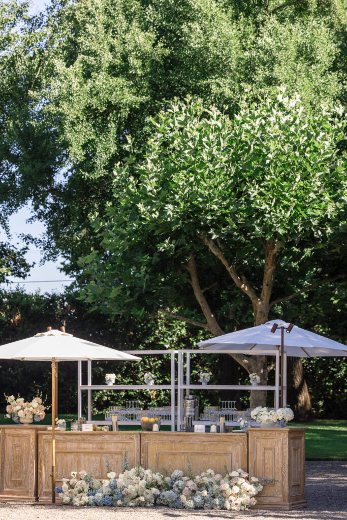 An elegant outdoor cocktail hour bar setup on the lawn at Beaulieu Gardens in Rutherford during a Napa Valley wedding, with wood bar structures, white umbrellas, and floral arrangements, photographed by Suzanne Karp Photography