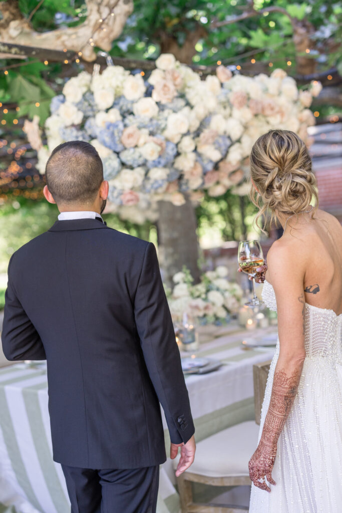 A bride and groom walk together toward the reception dinner beneath hanging floral installations at Beaulieu Gardens in Rutherford, the bride in a beaded strapless gown and the groom in a classic black tuxedo, photographed by Napa Valley wedding photographer Suzanne Karp