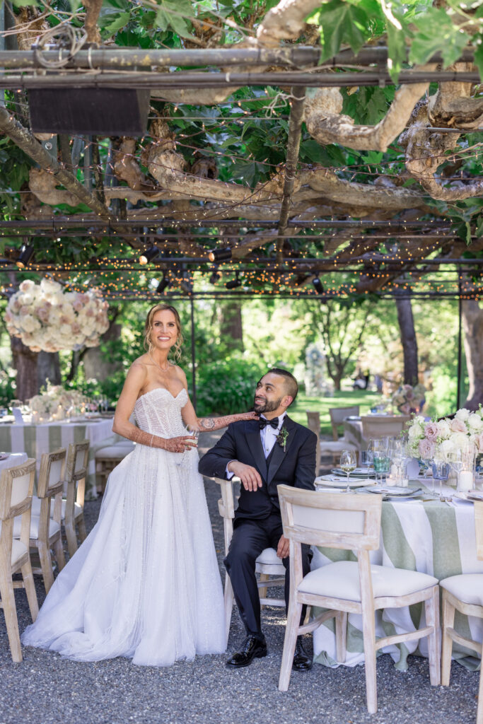 A bride and groom share a joyful moment together beneath the fig tree trellis at Beaulieu Gardens in Rutherford, surrounded by sage stripe linen tables and hanging floral installations during their Napa Valley wedding reception, photographed by Napa Valley wedding photographer Suzanne Karp