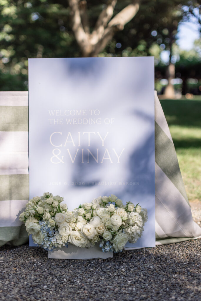 A frosted acrylic welcome sign reading Caity and Vinay surrounded by white garden rose and greenery arrangements on the lawn at Beaulieu Gardens in Rutherford during a Napa Valley wedding, photographed by Suzanne Karp Photography
