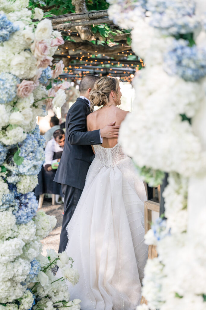 A bride and groom share their first dance framed by a lush floral arch of white hydrangea and blush roses beneath the fig tree trellis at Beaulieu Gardens in Rutherford during their Napa Valley wedding reception, photographed by Napa Valley wedding photographer Suzanne Karp