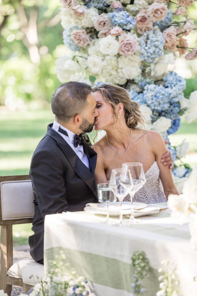 A bride and groom share a kiss at their sweetheart table surrounded by blue hydrangea and blush rose arrangements on sage stripe linens at Beaulieu Gardens in Rutherford during their Napa Valley wedding reception, photographed by Suzanne Karp Photography
