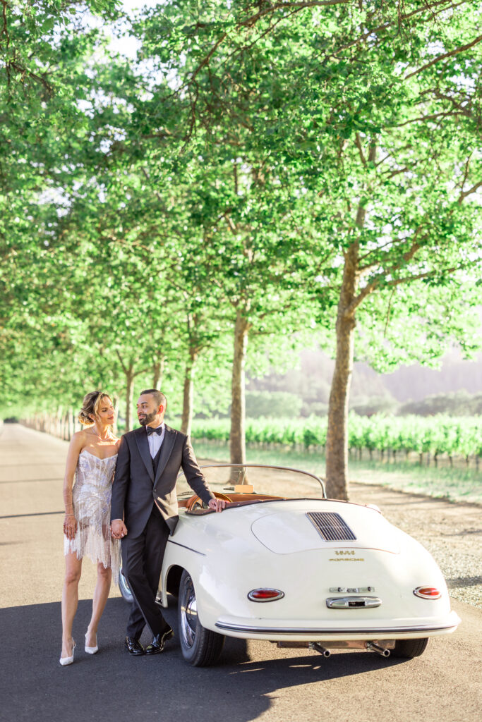 A bride and groom stand together beside a vintage white Porsche convertible on a tree lined vineyard road during their Napa Valley wedding portrait session, photographed by Napa Valley wedding photographer Suzanne Karp