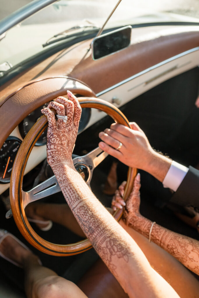 A close up detail of a bride's henna covered hands and gold bangles resting on the leather steering wheel of a vintage white Porsche convertible during a Napa Valley wedding, photographed by Napa Valley wedding photographer Suzanne Karp