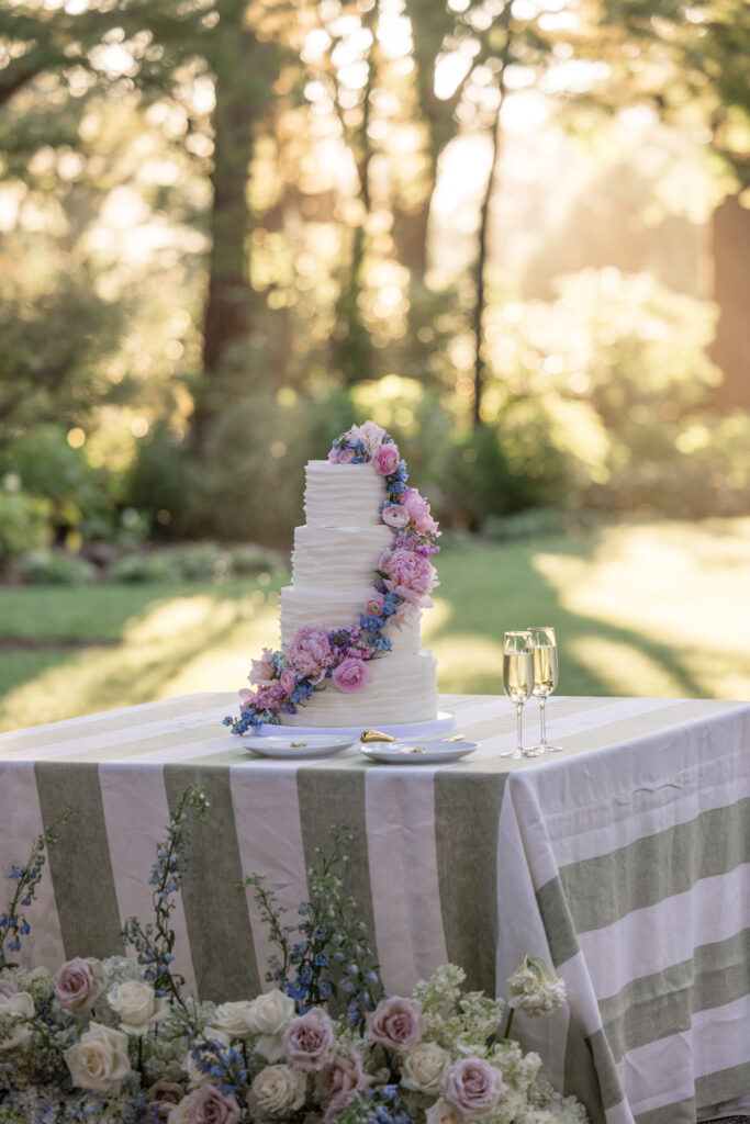A four tier white fondant wedding cake with a diagonal cascade of fresh flowers by Flour and Bloom displayed on a sage stripe linen table in golden afternoon light at Beaulieu Gardens in Rutherford, photographed by Napa Valley wedding photographer Suzanne Karp