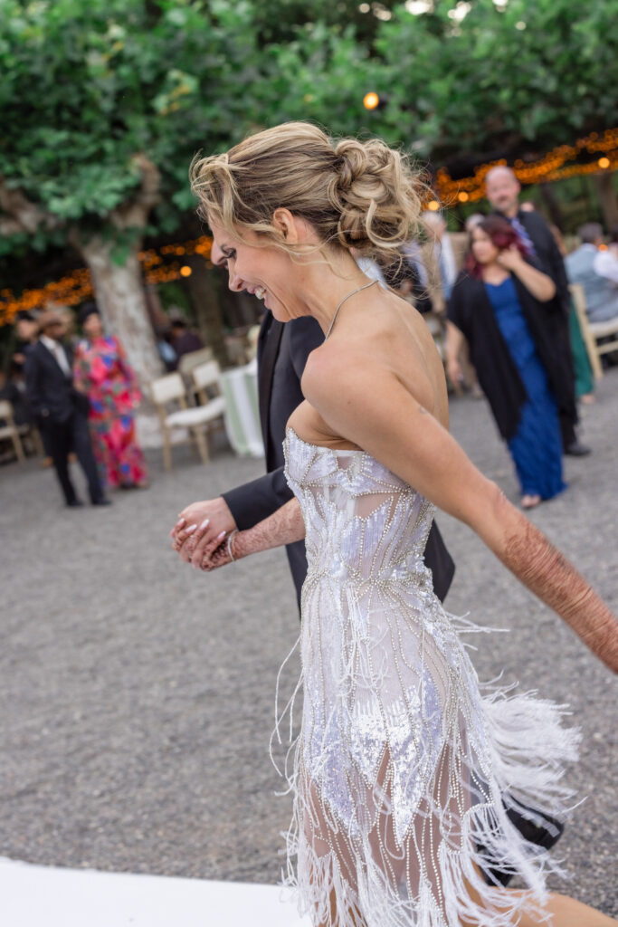 A bride laughs joyfully while dancing in a beaded fringe reception dress beneath the fig tree trellis at Beaulieu Gardens in Rutherford during her Napa Valley wedding reception, photographed by Napa Valley wedding photographer Suzanne Karp