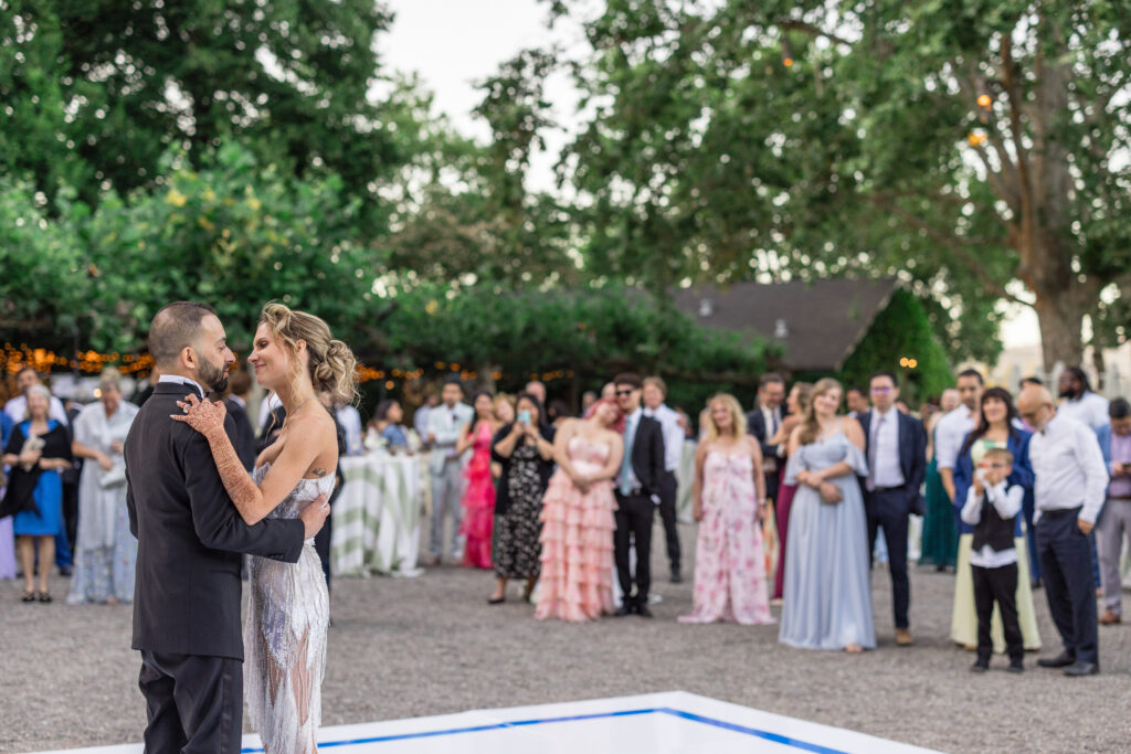A wide shot of a bride and groom sharing their first dance on an outdoor dance floor surrounded by their 200 wedding guests beneath mature oak trees at Beaulieu Gardens in Rutherford, Napa Valley, photographed by Napa Valley wedding photographer Suzanne Karp