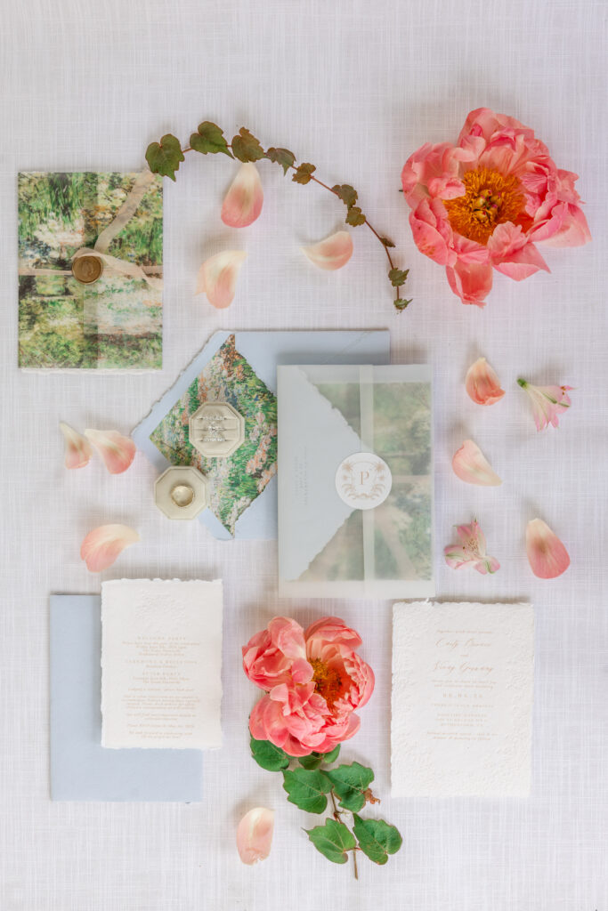 A wedding invitation suite from Tiny Details with deckled edge paper, a wax seal monogram, and scattered coral peonies and rose petals styled on a white linen surface, photographed by Napa Valley wedding photographer Suzanne Karp