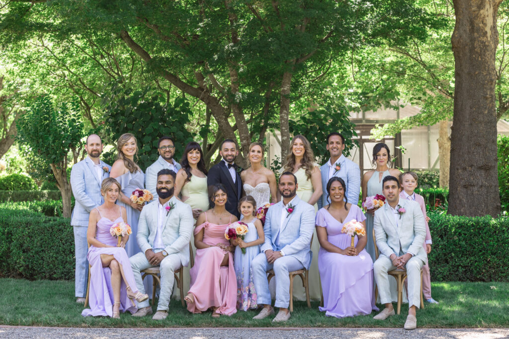 The full wedding party poses together beneath mature trees on the lawn at Beaulieu Gardens in Rutherford, the bridesmaids in soft lavender and pink gowns and groomsmen in light blue suits, photographed by Napa Valley wedding photographer Suzanne Karp