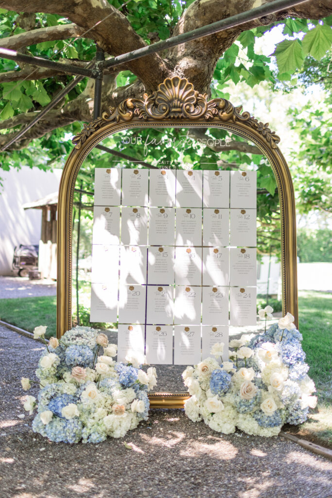 A gold ornate mirror displaying escort cards surrounded by white and blue hydrangea arrangements on the lawn at Beaulieu Gardens in Rutherford during a Napa Valley wedding, photographed by Suzanne Karp Photography