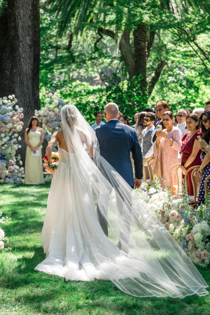 A bride in a beaded strapless gown with a cathedral veil trailing behind her walks down the floral lined aisle on her father's arm toward the ceremony arch at Beaulieu Gardens in Rutherford, photographed by Napa Valley wedding photographer Suzanne Karp