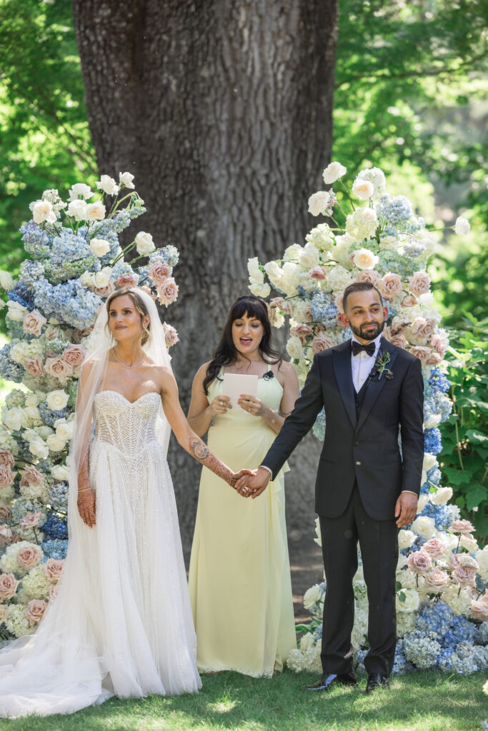 A bride and groom stand before a stunning asymmetrical flower arch of blue hydrangea, blush roses, and white blooms with their officiant during their Beaulieu Gardens wedding ceremony in Rutherford, Napa Valley, photographed by Suzanne Karp Photography