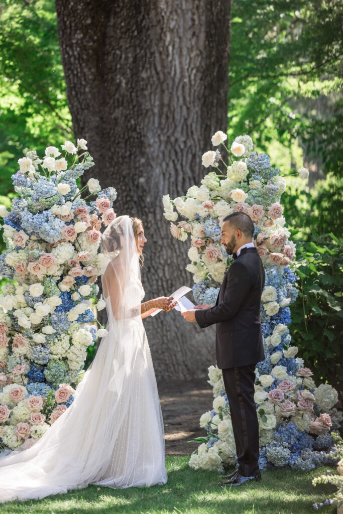 A bride reads her vows to her groom before a lush asymmetrical flower arch of blue hydrangea and blush roses at Beaulieu Gardens in Rutherford, photographed by Napa Valley wedding photographer Suzanne Karp