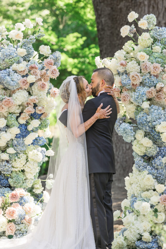 A bride and groom share their first kiss beneath a breathtaking asymmetrical flower arch of blue hydrangea, blush garden roses, and white blooms at Beaulieu Gardens in Rutherford, Napa Valley, photographed by Suzanne Karp Photography