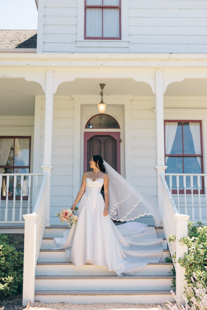 A bride in a strapless ballgown with a cathedral veil descends the steps of a white Victorian farmhouse surrounded by lavender during a Northern California wedding, photographed by Suzanne Karp Photography