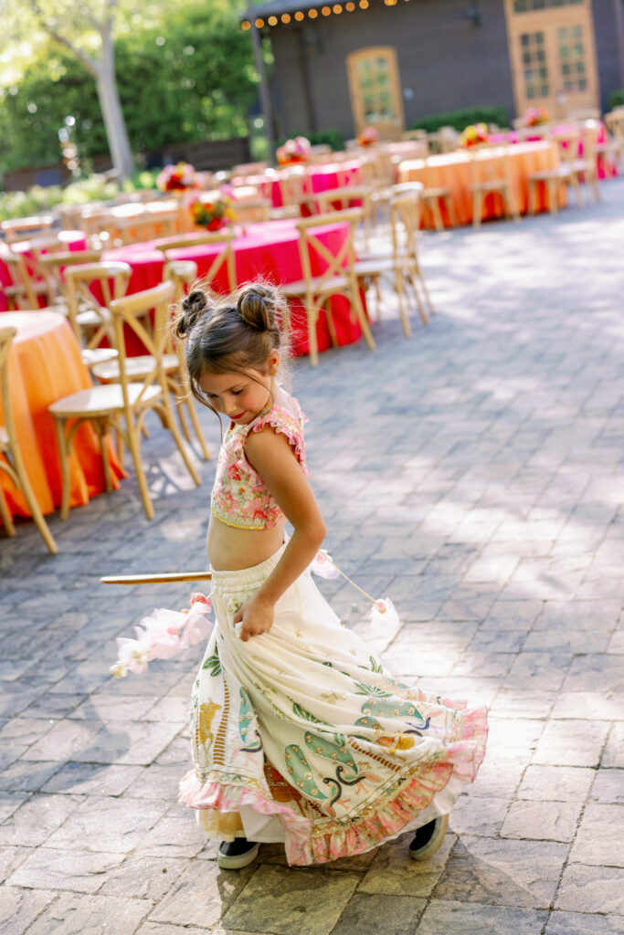 A young girl in an embroidered cream and floral lehenga twirls joyfully on the brick courtyard during the Sangeet celebration at Estate Yountville in Napa Valley, photographed by Napa Valley wedding photographer Suzanne Karp