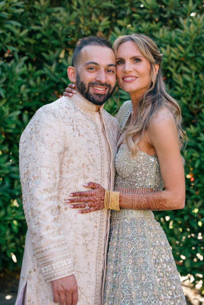 A bride and groom pose together in the lush garden at Beaulieu Gardens in Rutherford during their Napa Valley wedding, the bride in a silver embroidered lehenga with henna covered hands, photographed by Suzanne Karp Photography
